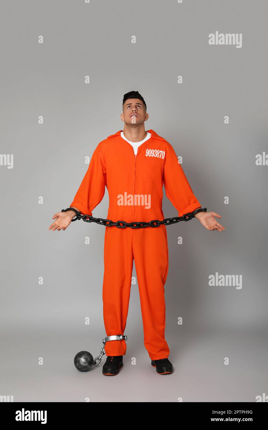 Gefangener in orangefarbenem Overall mit angeketteten Händen und Metallkugel auf grauem Hintergrund Stockfoto