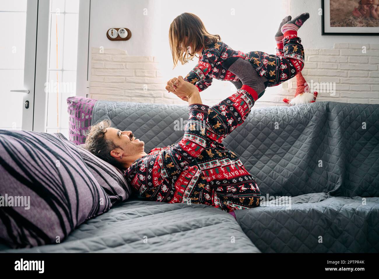 Dieses Bild zeigt einen Vater und eine Tochter im passenden Pyjama, wobei der Vater die Tochter zu Hause mit den Beinen auf dem Sofa hebt. Die Szene Stockfoto