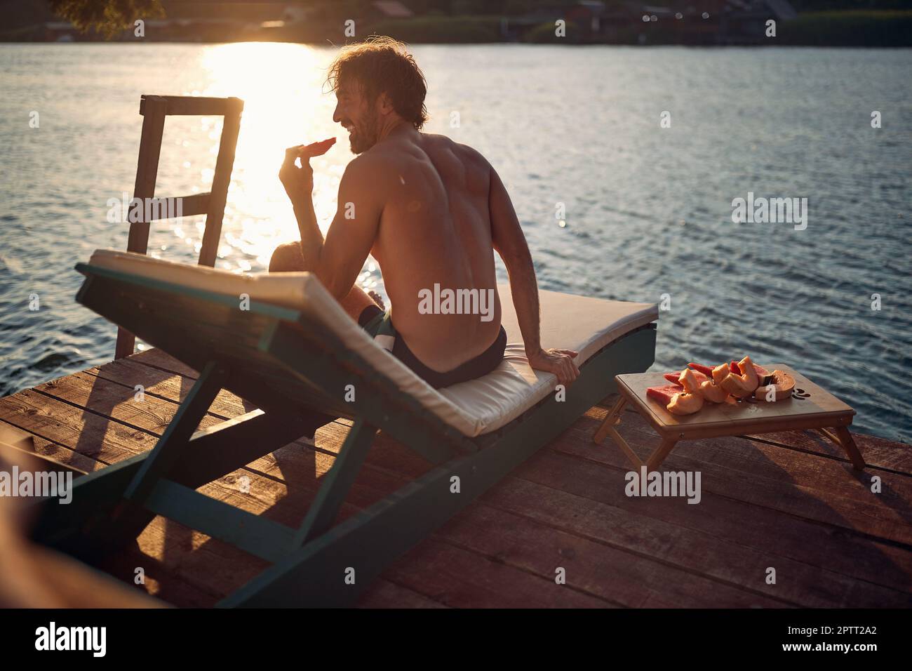 Ein junger Mann, der an einem wunderschönen Sommertag auf der Liege am Dock am Fluss Sonne und Obst genießt. Sommer, Fluss, Urlaub Stockfoto