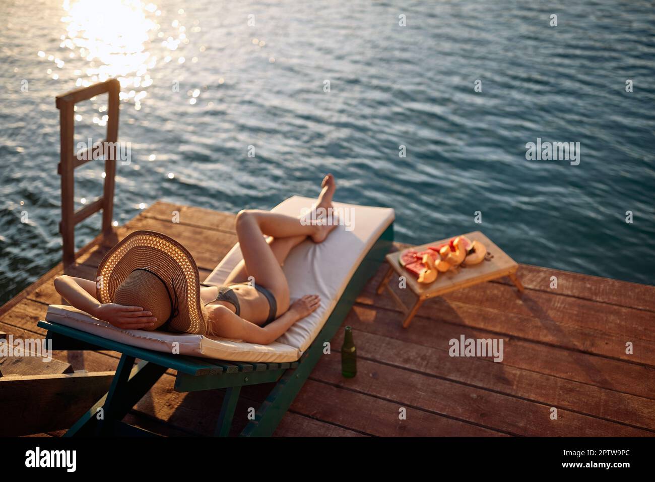 Mädchen entspannt am Dock. Junge attraktive Frau im Bikini auf einem Dock am Wasser während des Sommeruntergangs Stockfoto