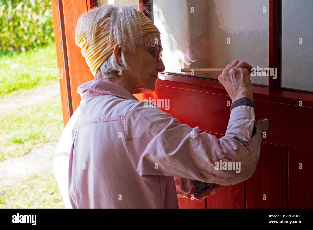 Ältere ältere ältere Frau, die Windwo-Rahmenelemente einer offenen Tür in Rot mit kleinem Pinsel und Farbe mit Overall malt Wales UK 2023 KATHY DEWITT Stockfoto