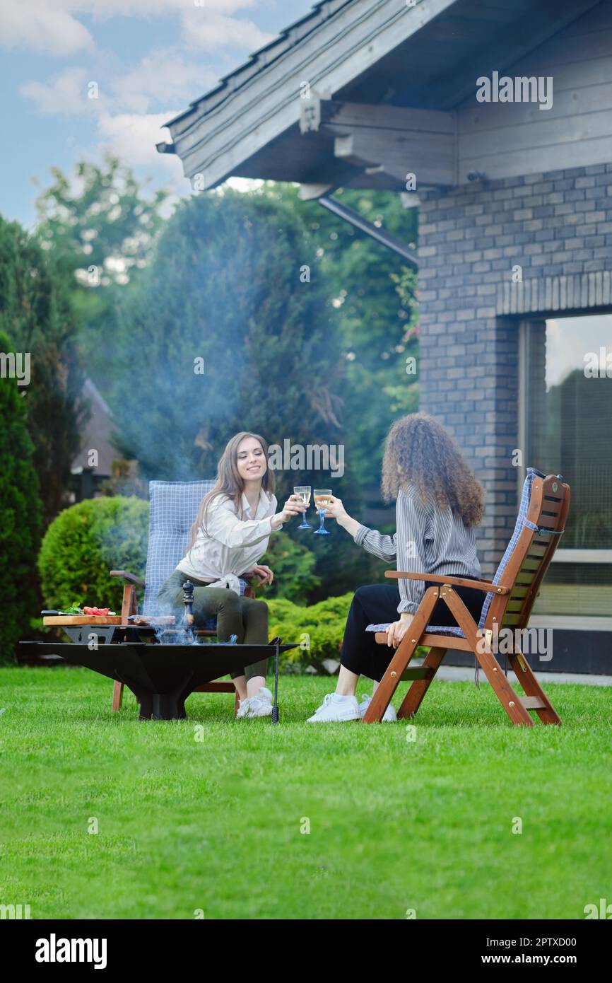 Zwei Freundinnen sitzen in der Nähe des Grills im Hinterhof und klirken Gläser mit Champagner Stockfoto