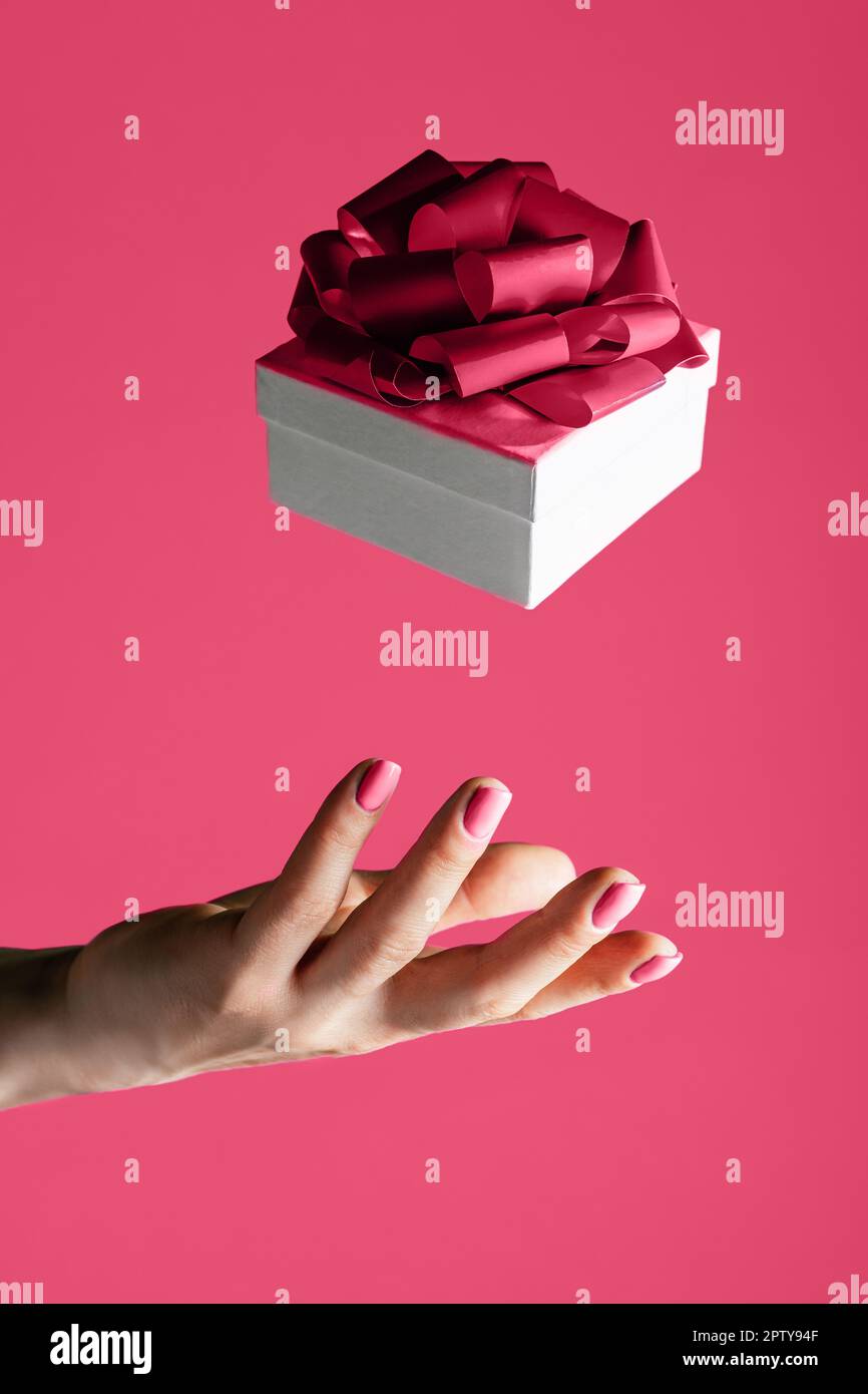 Weibliche Hand wirft in die Luft oder fängt weiße Geschenkbox mit Schleife auf dem Hintergrund von Color of the Year 2023 Viva Magenta. Schwebende Gabe. Vertikal Stockfoto