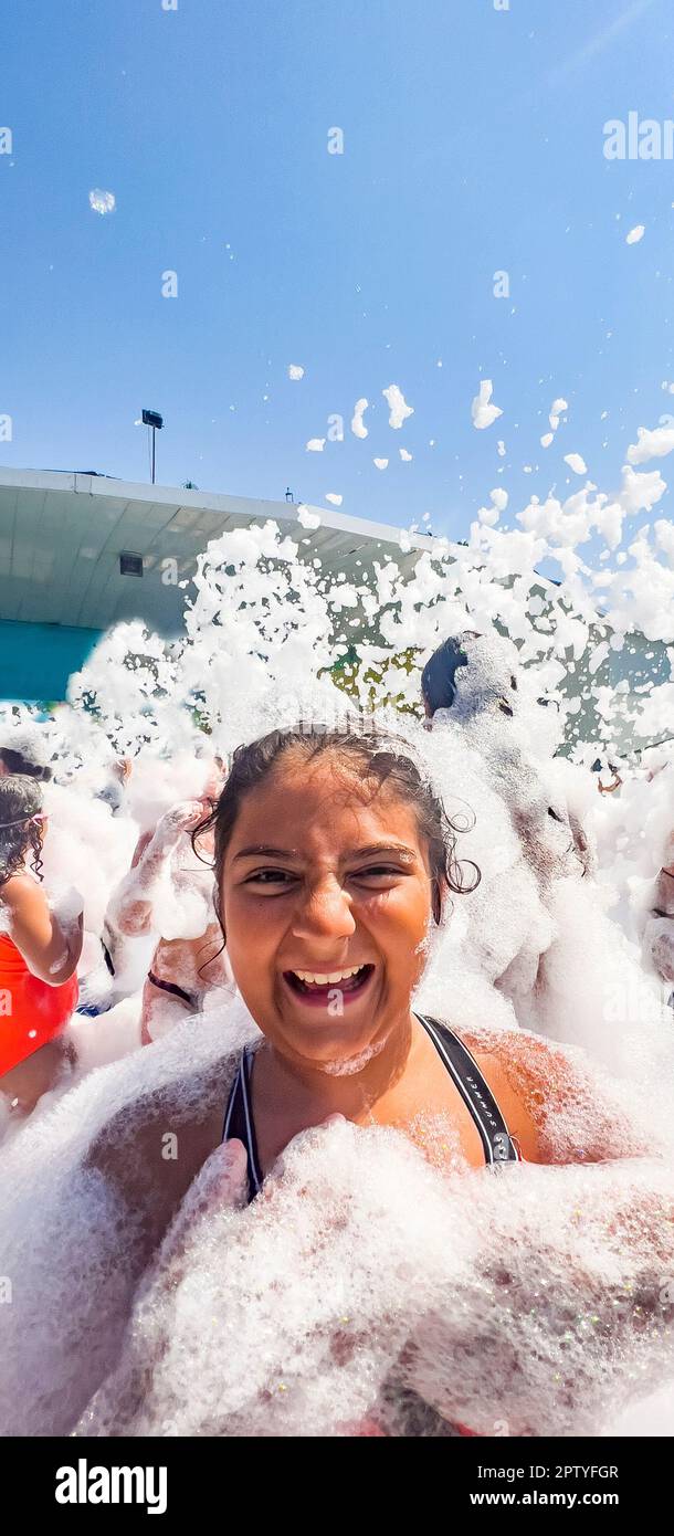 Fröhliches, lachendes Mädchen, mit Schaum bedeckt. Sommerkonzept: Verschwommene, nicht wiedererkennbare Leute, die auf einer Schaumbad-Party spielen. Kinder tanzen auf der Tanzfläche Stockfoto