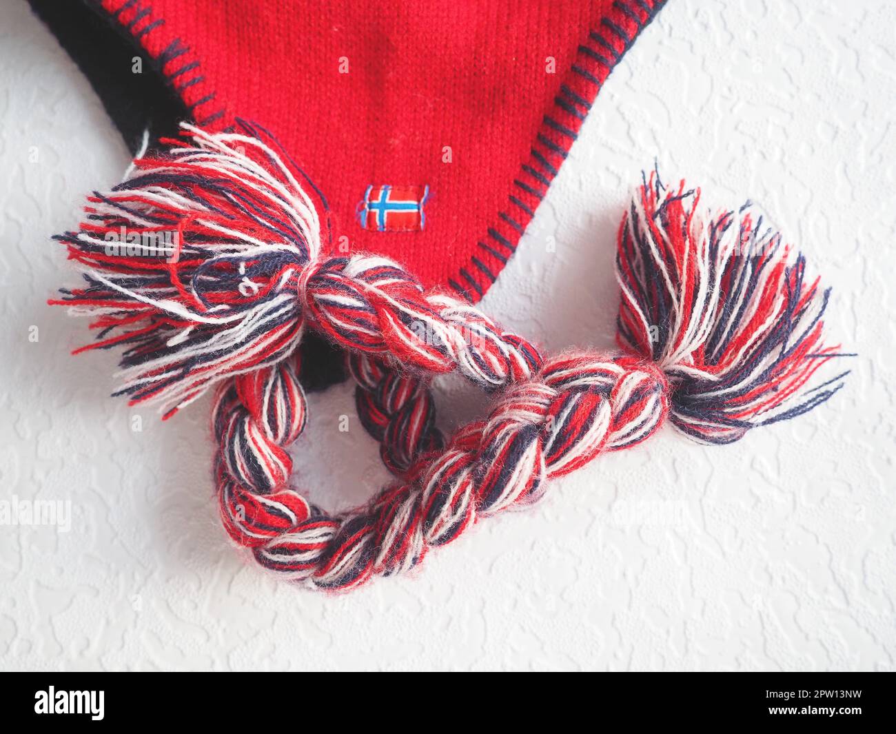 Kappe mit Ohrenklappen im norwegischen Stil. Gestrickter Modeartikel mit Krawatten und Bommeltaschen. Rotes Garn mit schwarz-weißen Fäden. Aufgenähte Minifahne Stockfoto