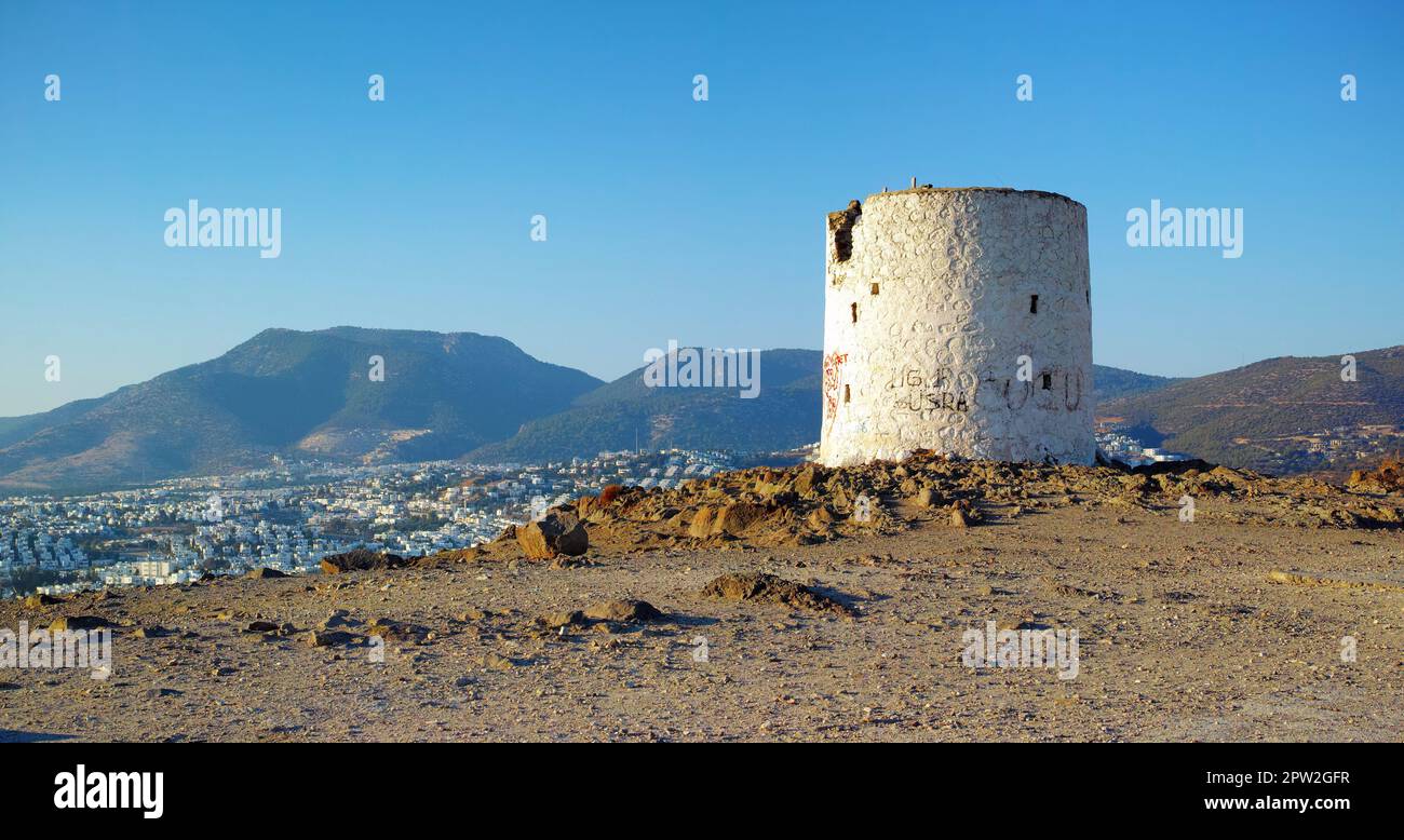 Ruinen einer kaputten Windmühle auf einem Hügel mit Blick auf eine Stadt, die von Bergen umgeben ist. Zerbröckelnde Mauern des verlassenen Leuchtturmturms. Ein verwittertes rundes Weiß Stockfoto