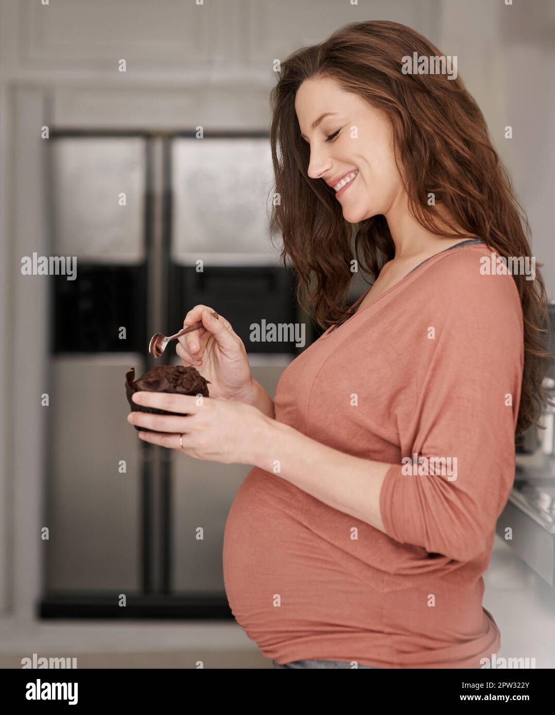 Sich ihrem Schwangerschaftsverlangen hingeben. Eine schwangere Frau, die einen Cupcake in der Küche isst Stockfoto