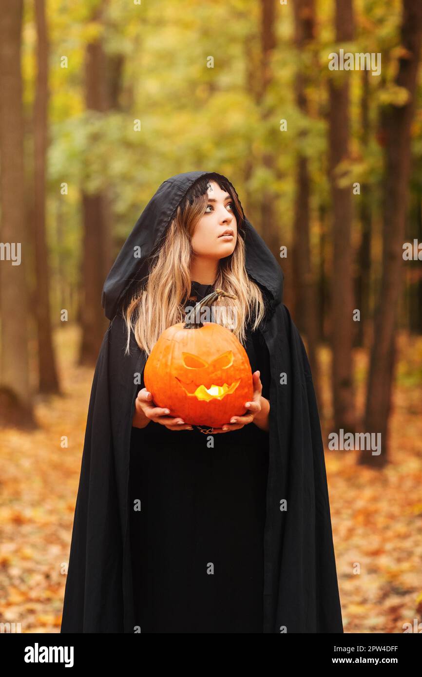 Junge Frau in schwarzem Umhang mit gruseligen Jack o Laterne Und auf den Halloween-Tag im Herbstwald wegschauen Stockfoto