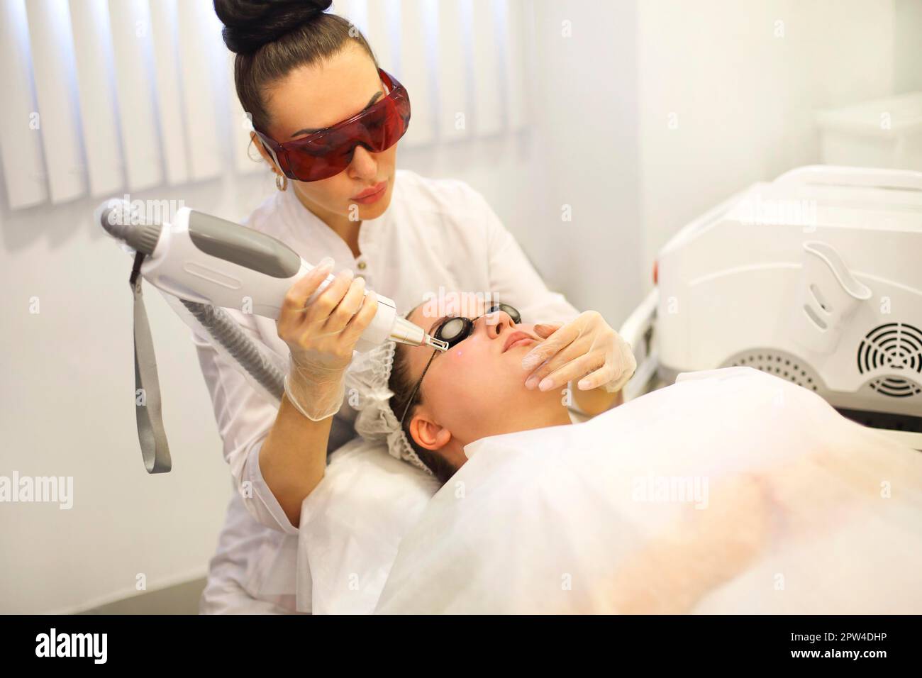 Fokussierte Kosmetikerin, die Laser-Resurfacing an den Kunden in einer Brille macht Im Büro auf der Couch liegen Stockfoto
