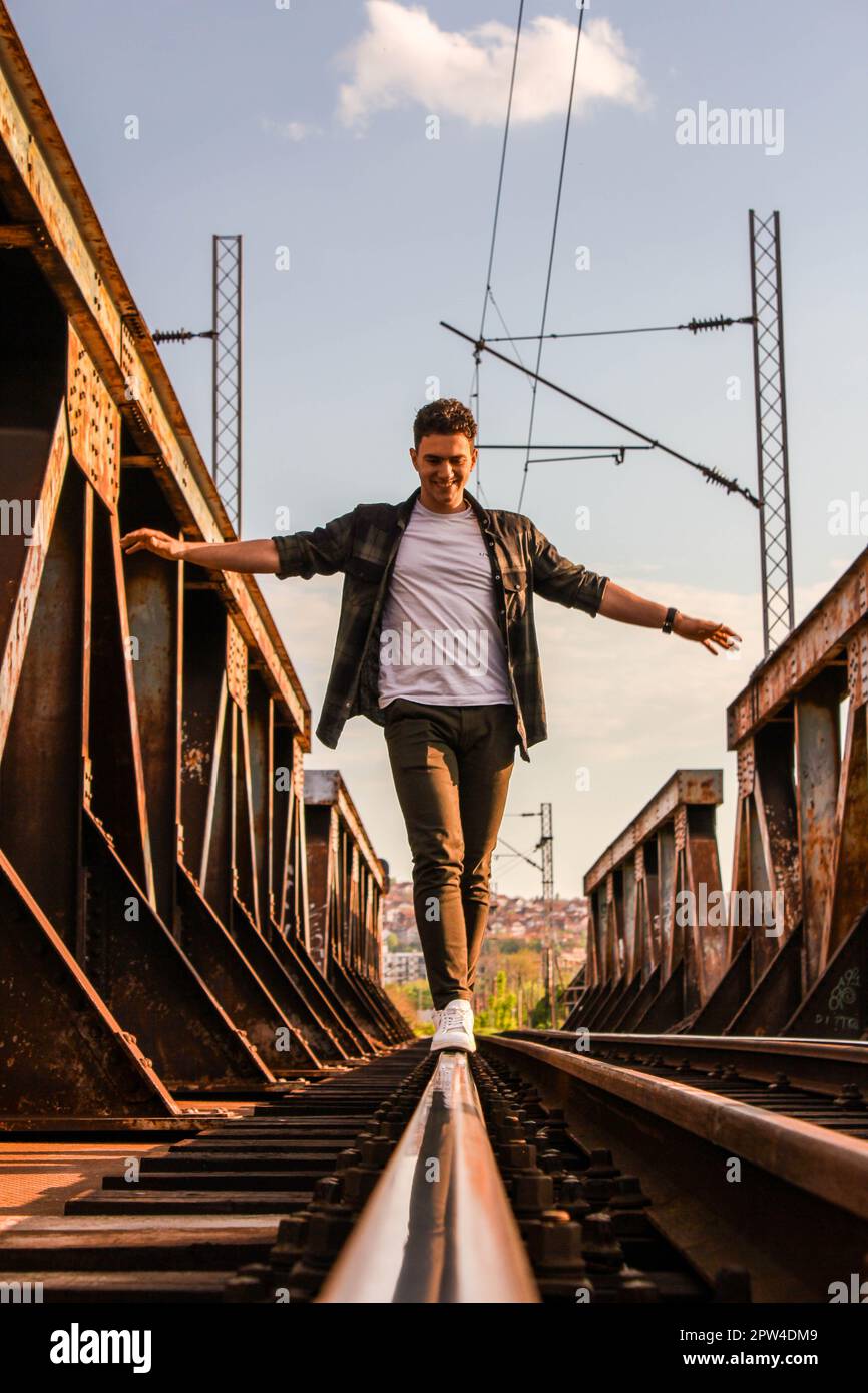 Ein kaukasischer Teenager, der vorsichtig auf Bahngleisen auf einer rostigen Eisenbahnbrücke mit einer verschwommenen Stadt im Hintergrund spaziert Stockfoto