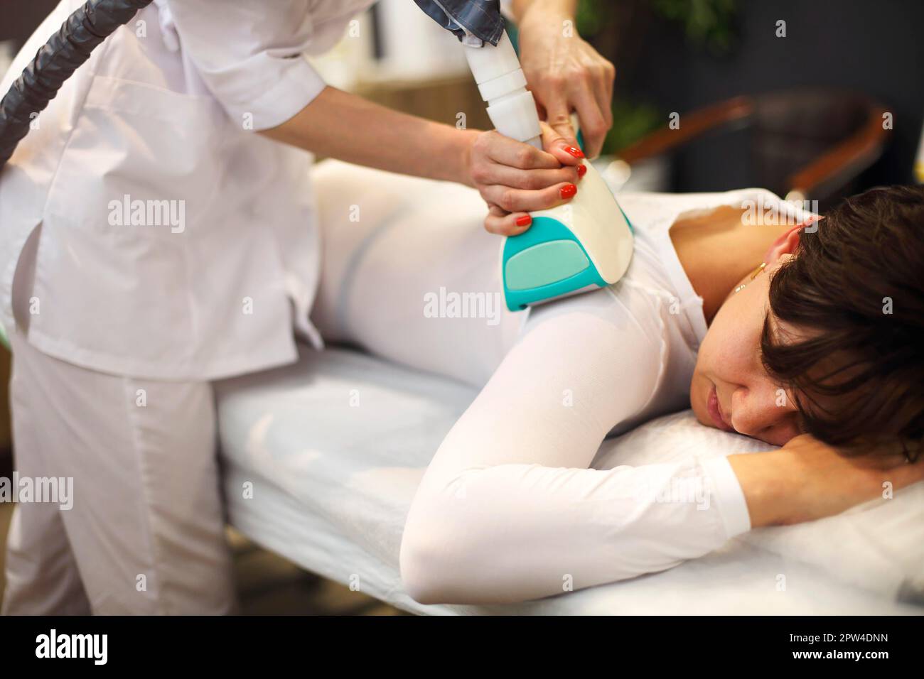 Gesichtslose Kosmetikerin, die Flüssiggas-Schlankheits-Apparate am weiblichen Körper verwendet Klient in weißem Anzug, der auf der Couch liegt Stockfoto