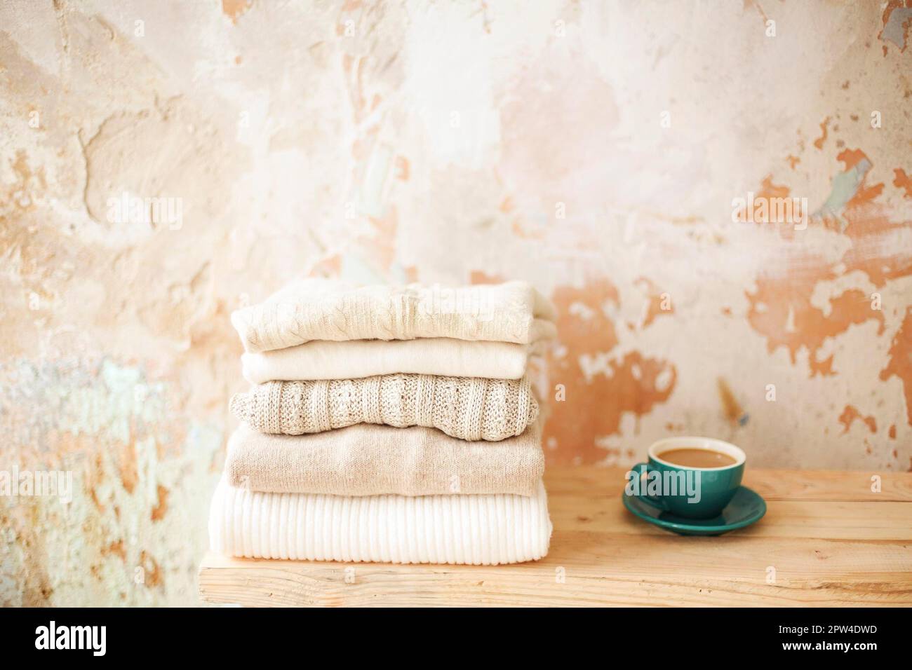 Tasse frischen Kaffee auf einem Holzregal in der Nähe eines Stapels warmer gestrickter Kleidungsstücke an der schäbigen Wand Stockfoto