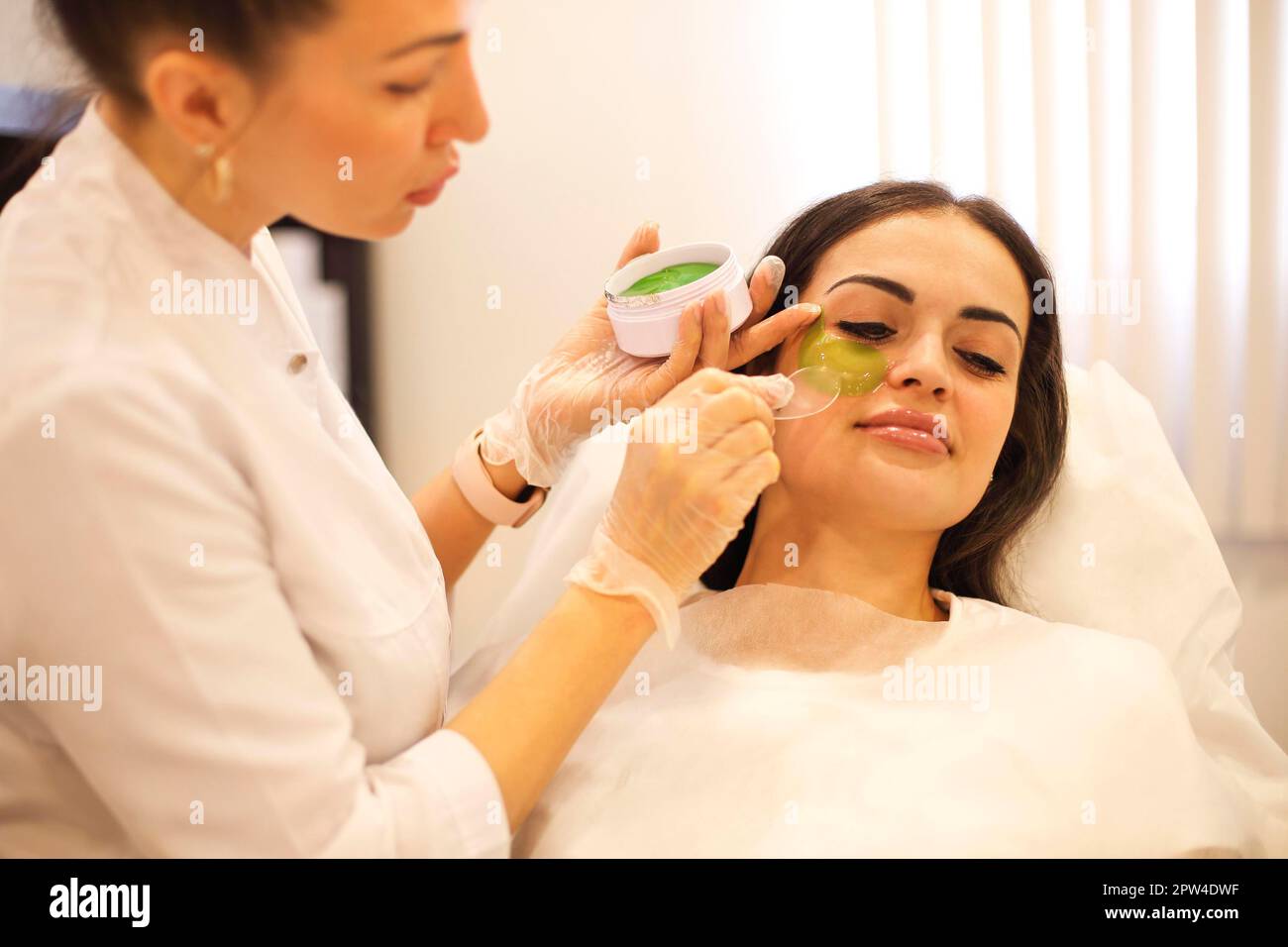 Professionelle Kosmetikerin setzt grüne Flecken auf den ruhigen Bereich unter den Augen Weibliche Besuch Schönheitsklinik Stockfoto