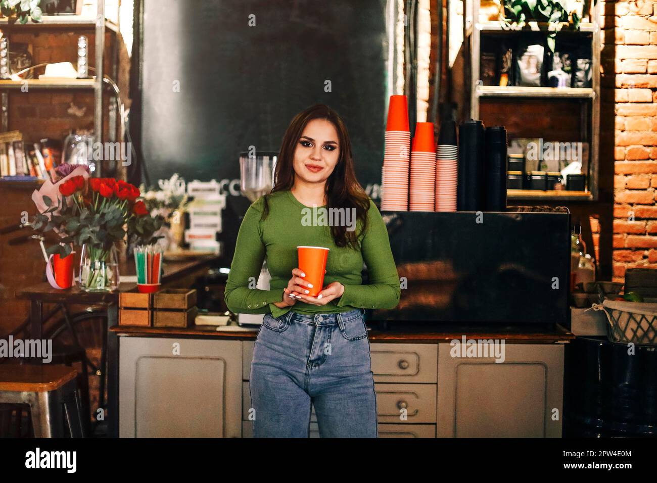 Junge Frau mit einer Tasse Kaffee schaut in die Kamera, während sie in der Nähe der Theke in einem gemütlichen Café steht Stockfoto