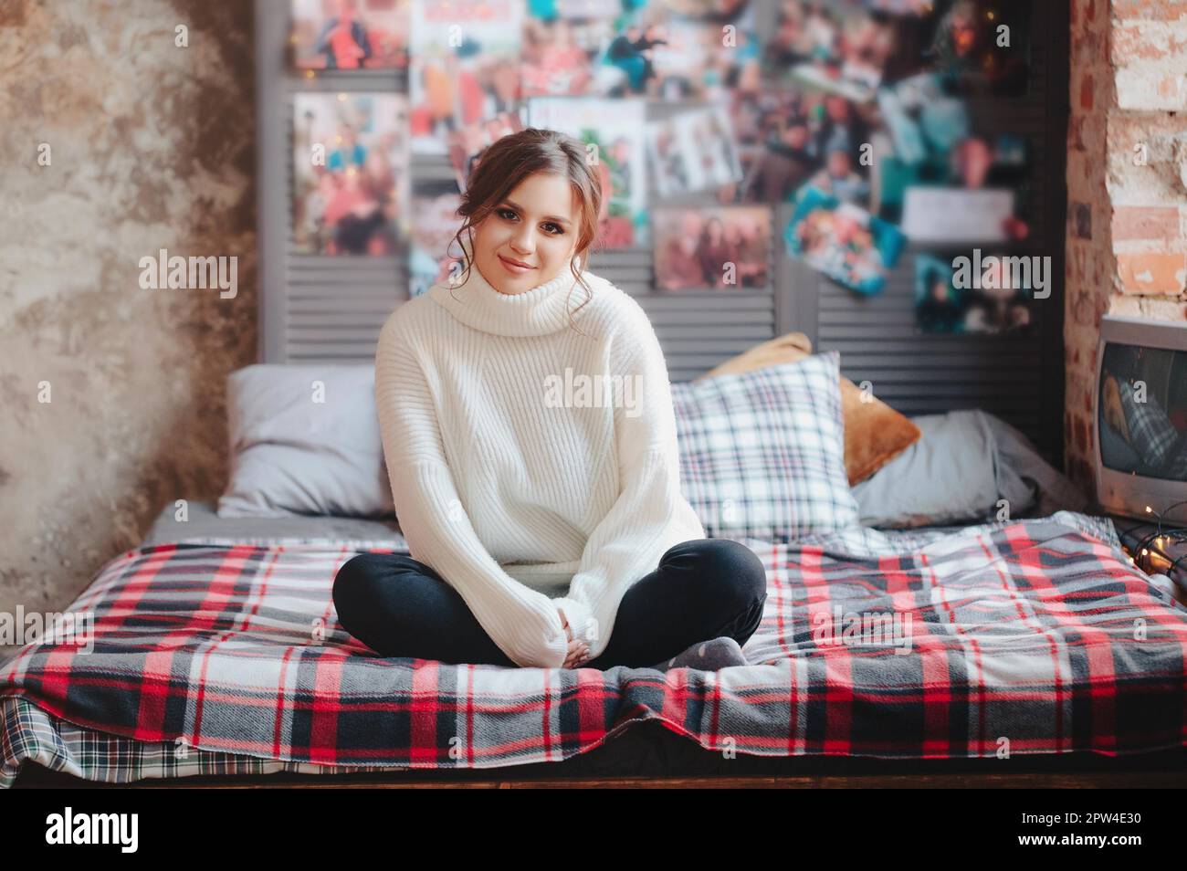 Junge Frau in warmem Strickpullover, die Kamera anschaut, während sie auf einem bequemen Bett mit kariertem Kariert an der Wand mit Vintage-Postern zu Hause sitzt Stockfoto