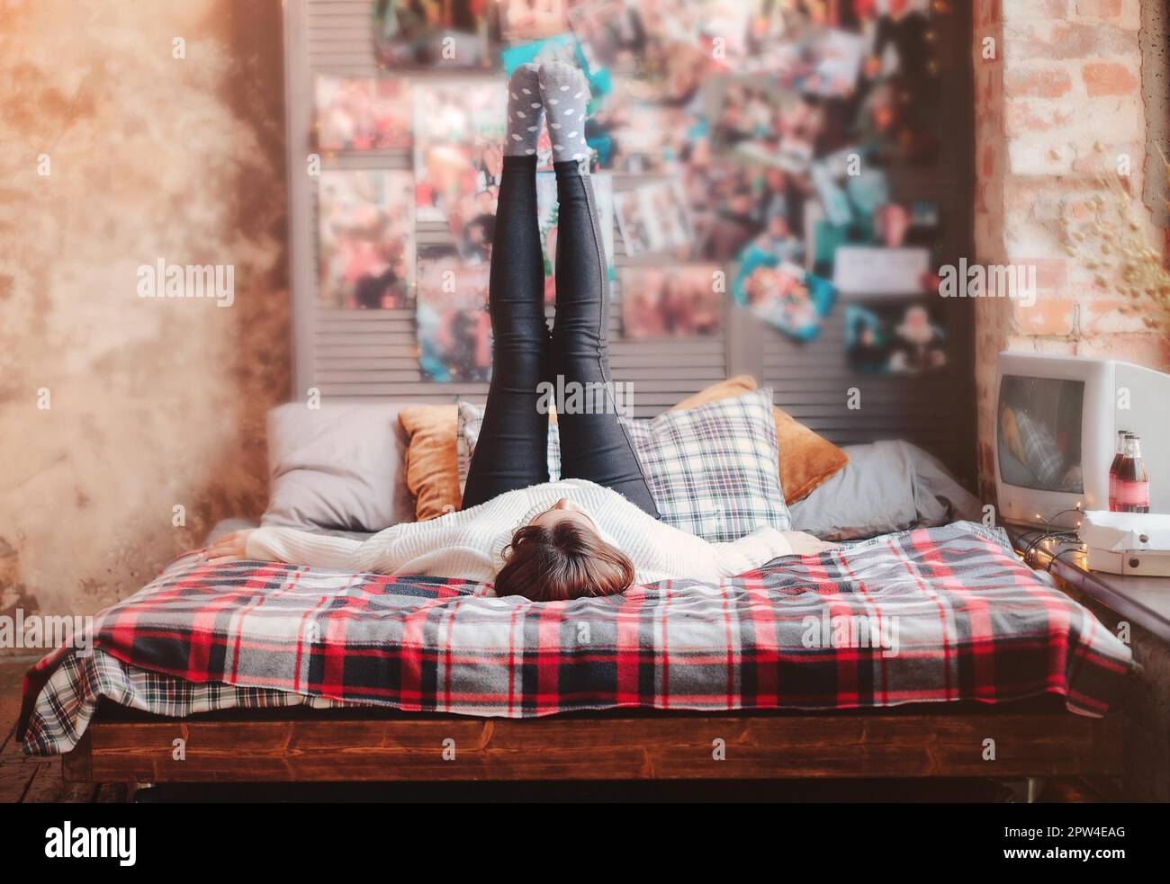 Junge Frau im warmen Strickpullover auf komfortablem Bett mit kariertem Kariert an der Wand mit Vintage-Postern zu Hause Stockfoto