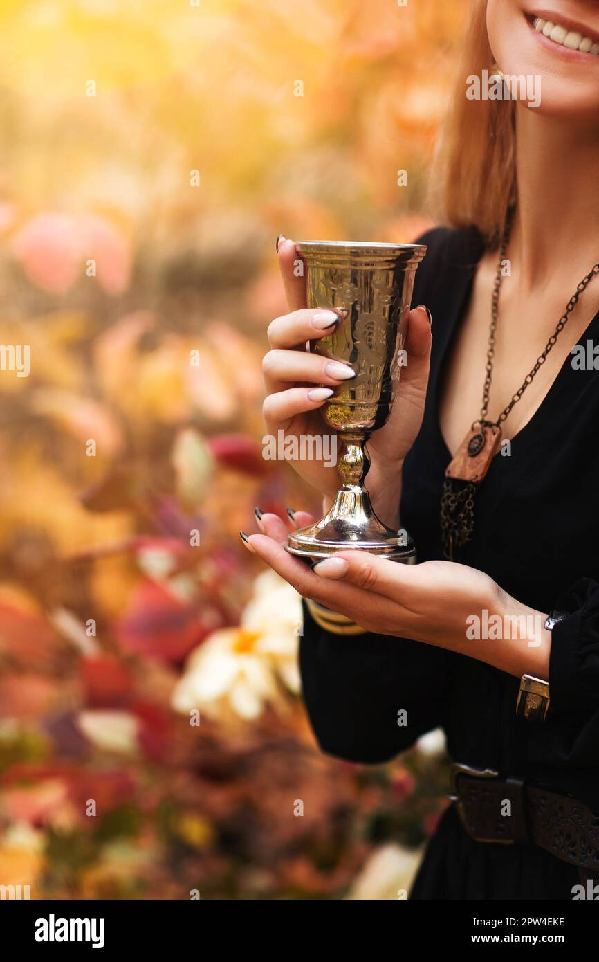 Kurze Aufnahme einer jungen, fröhlichen Frau in schwarzem Kleid, die altmodisches Metallglas in den Händen hält, während sie im Herbstpark mit bunten Blättern steht Stockfoto