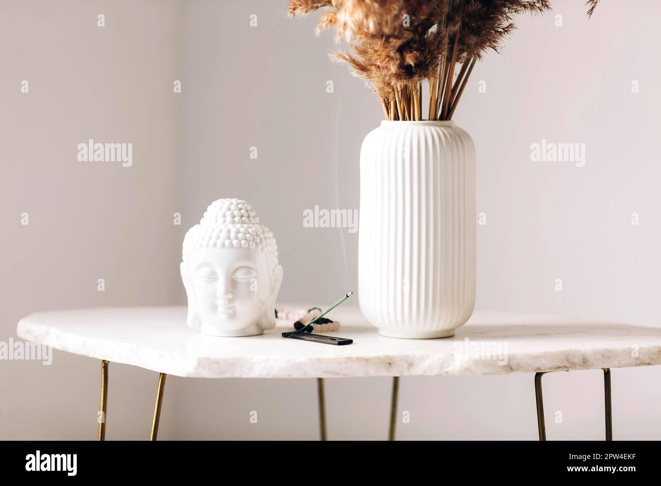 Kleiner Marmortisch mit Dekorationen, weiße Vase mit trockenen Blumen, kleine buddha-Statue und brennender Räucherstäbchen auf einem Stand in moderner Einfachheit Stockfoto