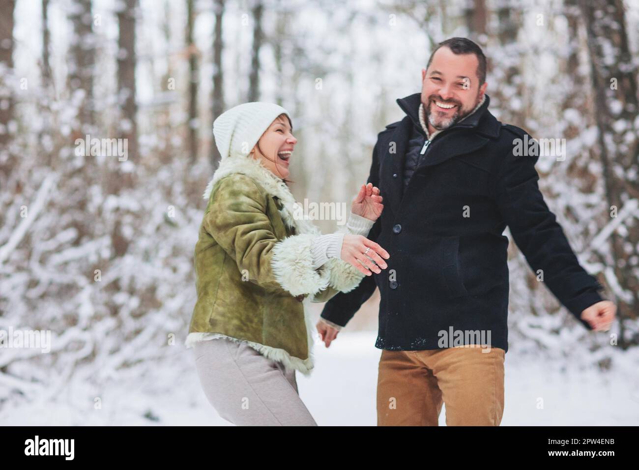 Schönes, glückliches Familienpaar mittleren Alters, das im Winter zusammen Spaß im Freien hat, einen Spaziergang durch den verschneiten Wald mit einem Lächeln, angezogen Stockfoto