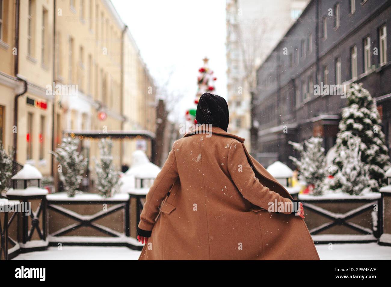 Junge positive Frau mit Außenseite in beigefarbenem Mantel und schwarzem Schal auf dem Kopf, zitternd von kaltem Winterwetter vor dem Hintergrund von Bäumen bedeckt mit Stockfoto