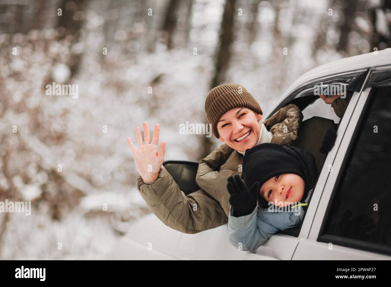 Fröhliche Erwachsene Frau und kleiner Junge in Oberbekleidung, die in die Kamera schauen und mit den Händen schwenken, während sie aus dem Fahrzeugfenster auf einen unscharfen Hintergrund schauen Stockfoto