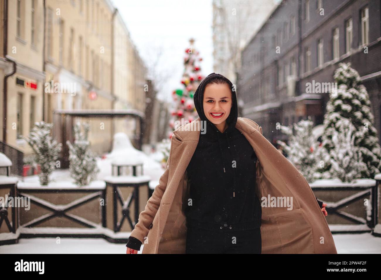 Junge positive Frau mit lustigem Lächeln im Gesicht steht draußen in beigefarbenem Mantel und schwarzem Schal auf dem Kopf und zittert vor kaltem Winterwetter Stockfoto