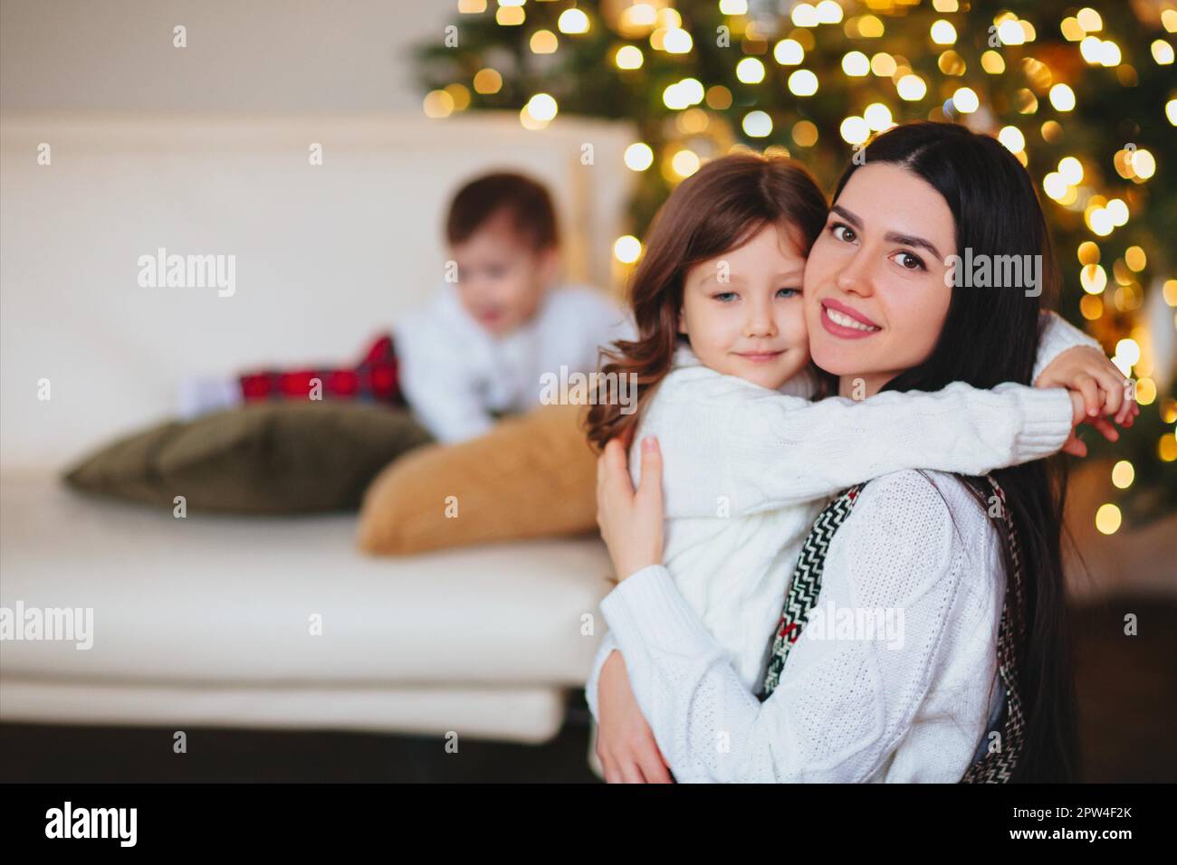 Junge Frau mit Kindern, die sich während der Weihnachtsfeier zu Hause in der Nähe von Nadelbäumen umarmt Stockfoto