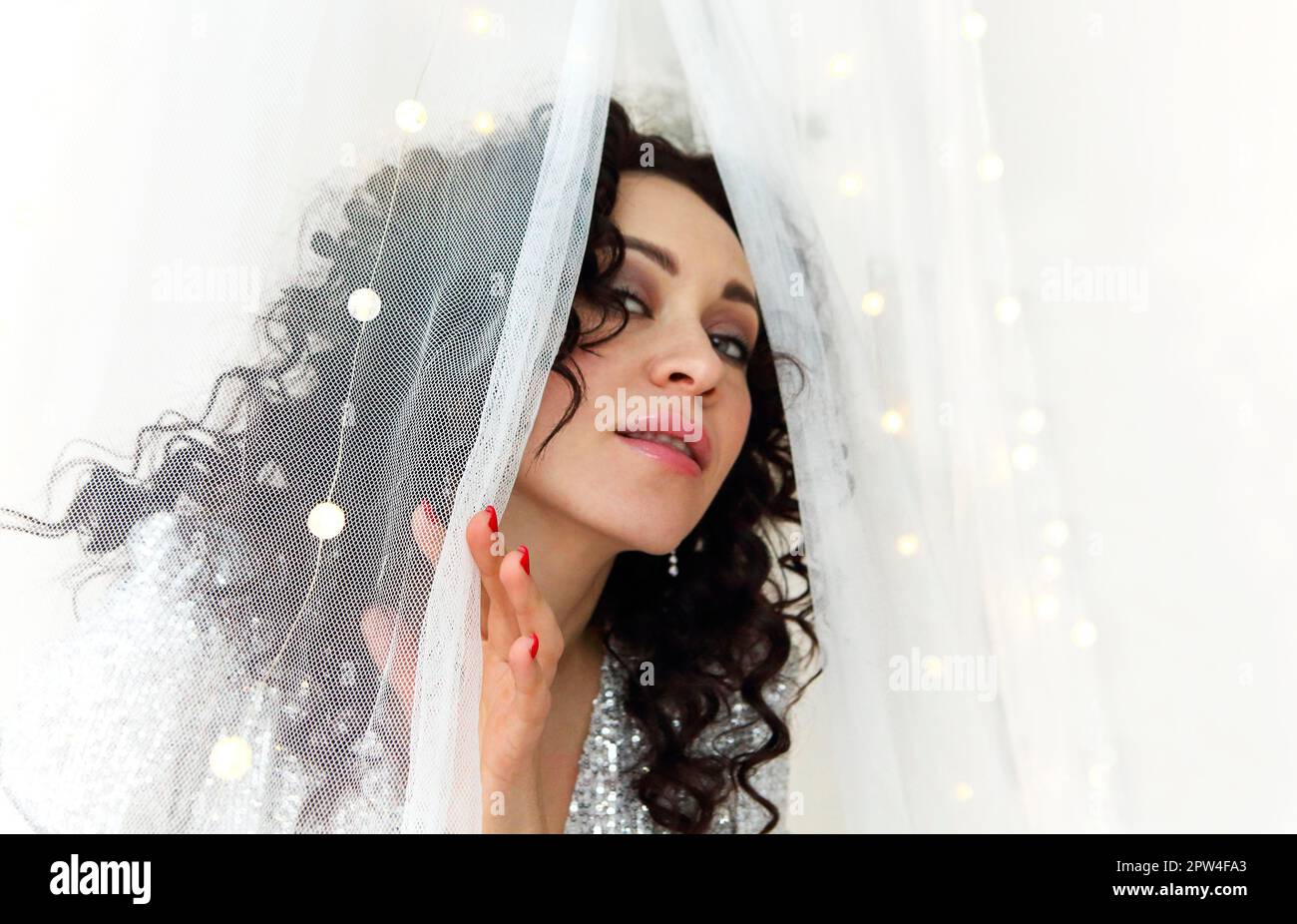 Nahaufnahme einer jungen, sinnlich attraktiven Frau mit lockigem Haar, die festliches Make-up trägt und hinter einen weißen Vorhang blickt, der mit Xmas LED dekoriert ist Stockfoto