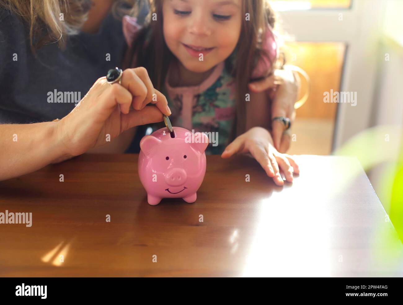 Kleines Mädchen, das Münze in Sparschwein legt, während es mit der Mutter zu Hause am Tisch sitzt, eine Frau, die dem Kind beibringt, Geld klug zu budgetierenund die Grundlagen erklärt Stockfoto