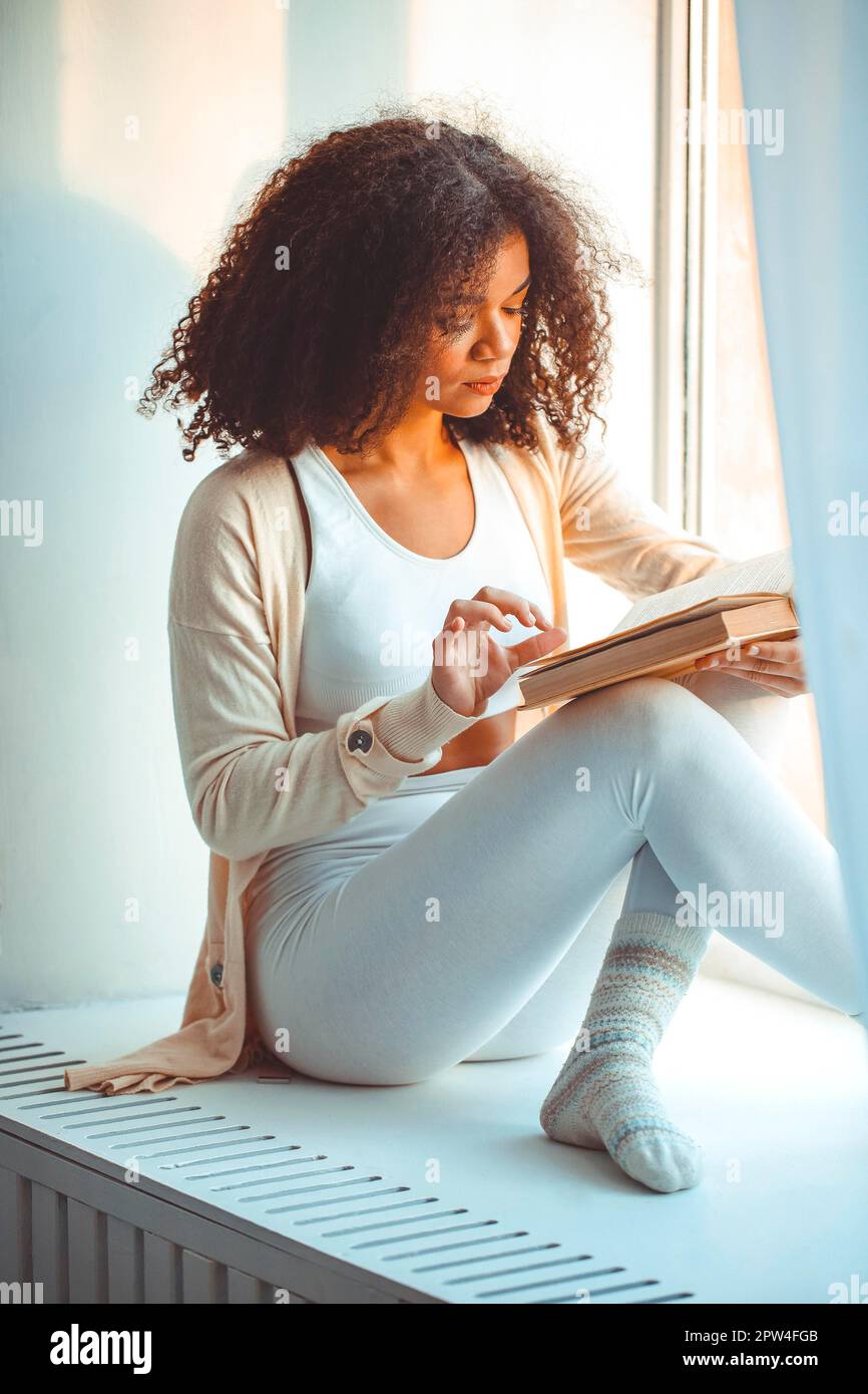 Mein Hobby ist Lesen. Ruhige, entspannte junge afroamerikanische Frau, die zu Hause auf dem Fensterbrett ein Buch liest, schwarzes, lockiges Mädchen am Fenster, das sich entspannt Stockfoto