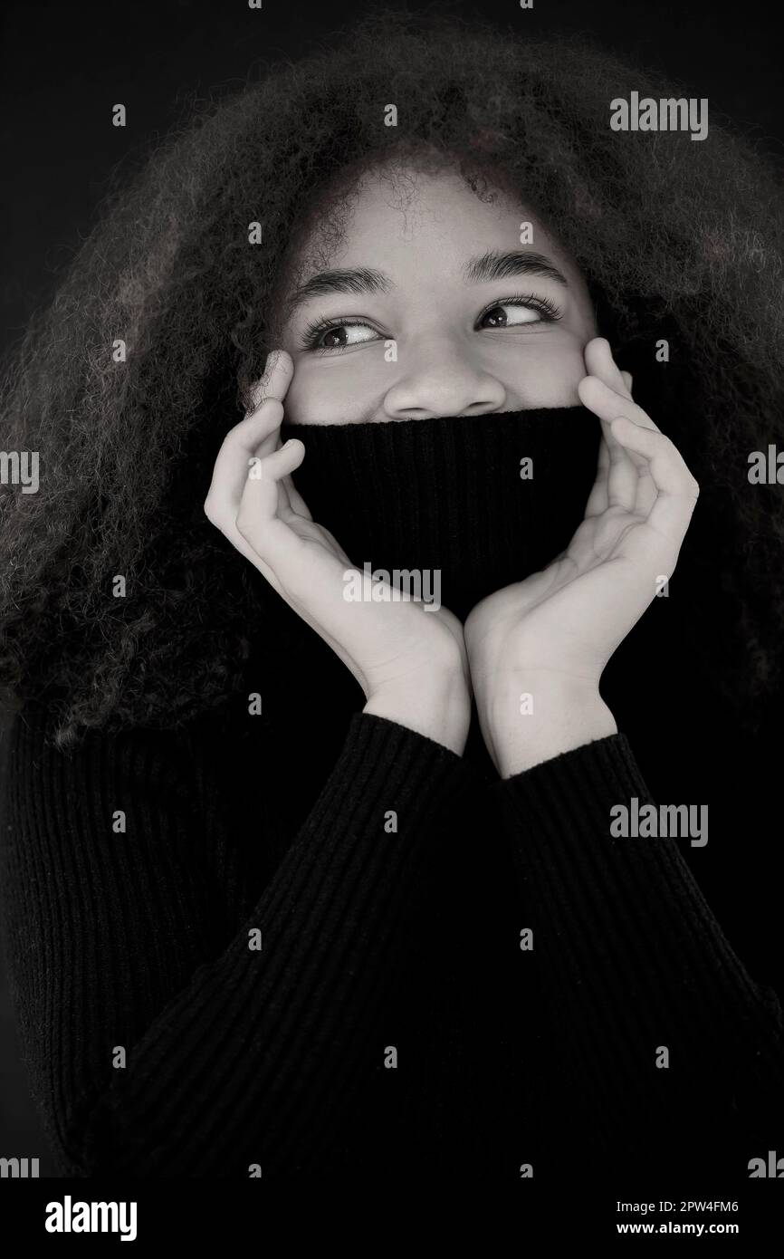 Erfreut afroamerikanische lockige Frau, die Augen schließt und Gesicht mit warmem schwarzen Pulloverkragen bedeckt, isoliert auf dunklem Studiohintergrund, lächelnd weiblich Stockfoto