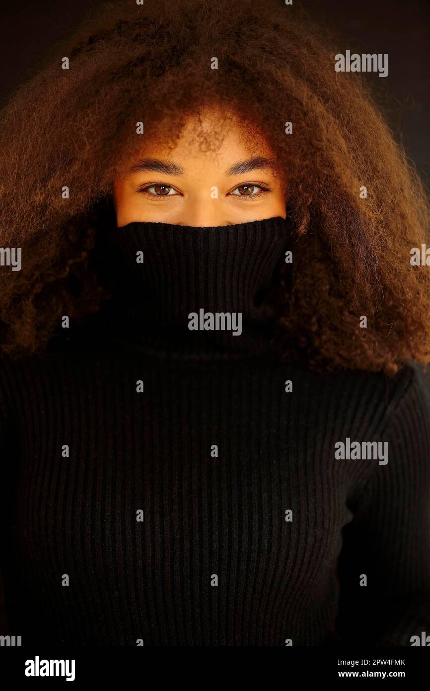 Erfreut afroamerikanische lockige Frau, die Augen schließt und Gesicht mit warmem schwarzen Pulloverkragen bedeckt, isoliert auf dunklem Studiohintergrund, lächelnd weiblich Stockfoto