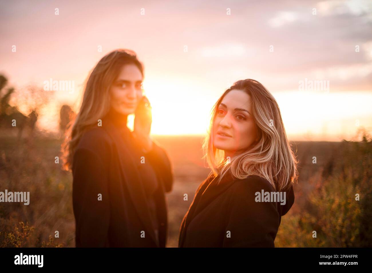 Zwei fröhliche Frauen, die Händchen halten, gehen bei Sonnenuntergang durch das Herbstfeld und lächeln, sorgenfreie weibliche beste Freunde, die den Abend genießen Stockfoto