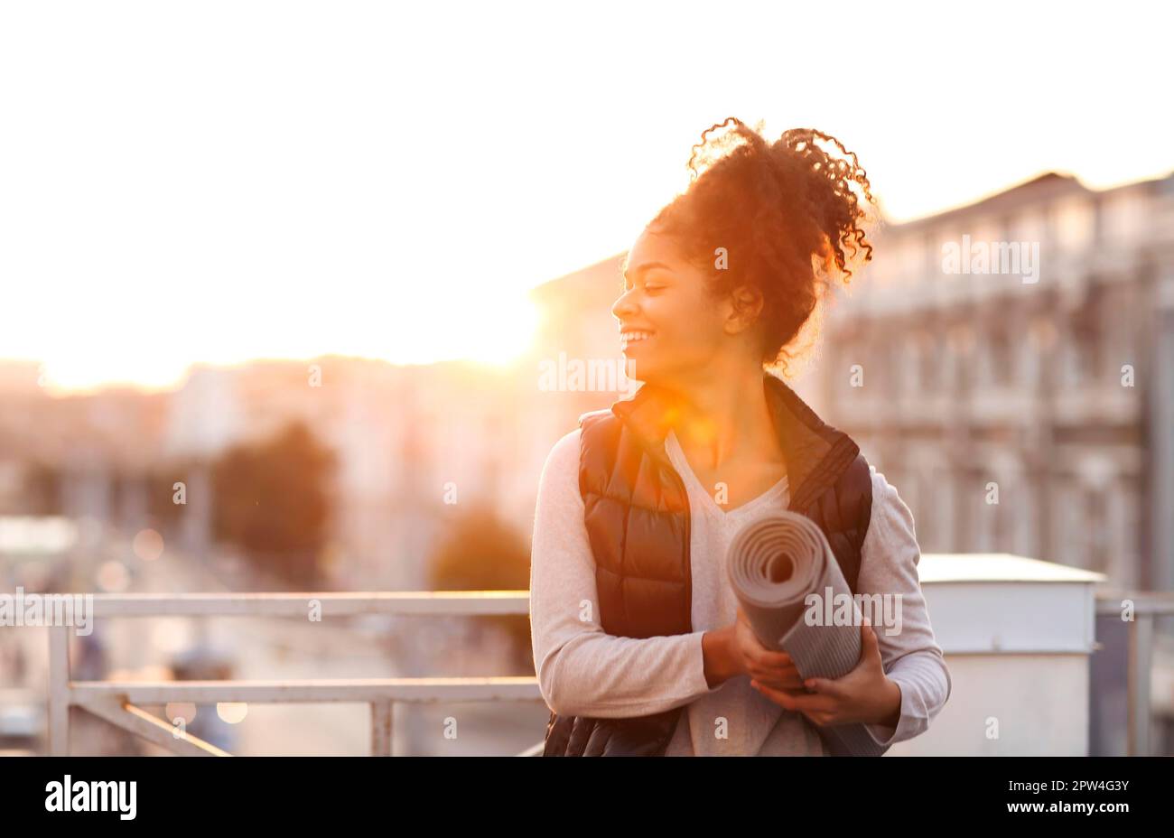 Eine lange, sportliche afroamerikanische Frau mit Yoga-Matte in den Händen auf dem Dach vor dem morgendlichen Outdoor-Workout, fit für gemischte Rennen Stockfoto