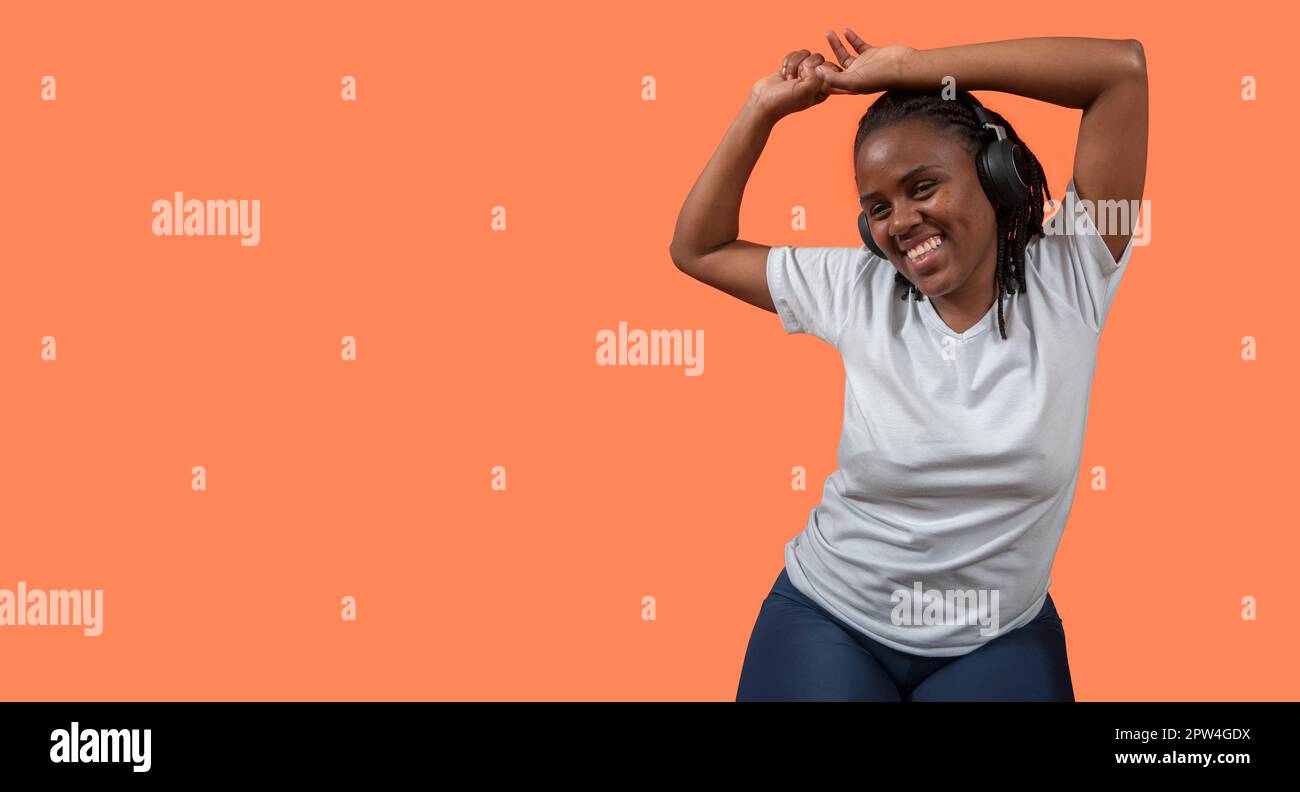 Porträt einer glücklichen jungen Afrikanerin, die mit Kopfhörern tanzt und Musik hört, Arme hoch, in einem weißen T-Shirt und einer blauen Hose gegen Orange gekleidet Stockfoto