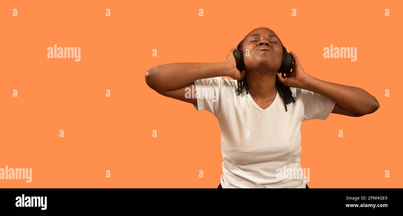 Porträt einer glücklichen jungen afrikanischen Frau, die Musik mit Kopfhörern hört, die Augen geschlossen hat, den Kopf nach oben hält und die Kopfhörer mit den Händen hält, in weißen T-Shirts Stockfoto