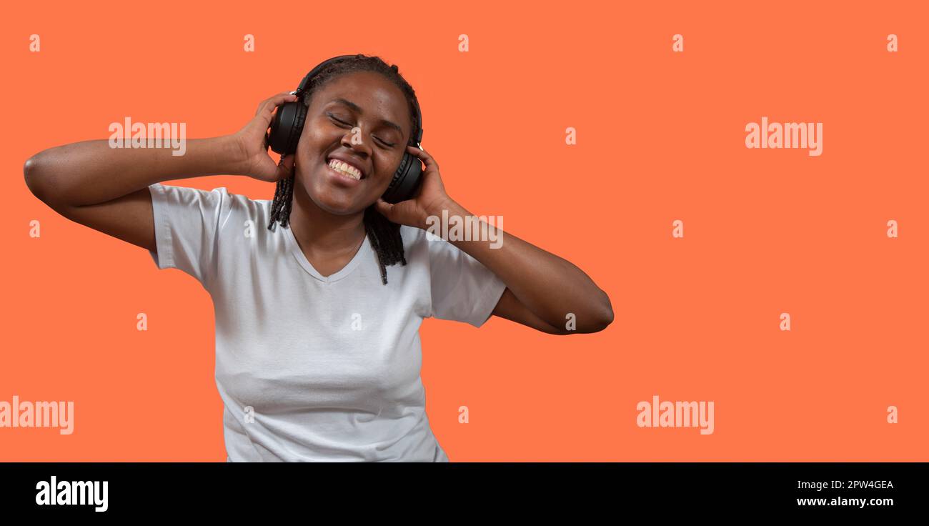 Porträt einer glücklichen jungen afrikanischen Frau, die Musik über Kopfhörer mit einem zahnigen Lächeln hört, Kopfhörer mit Händen hält, gekleidet in weißer T-Shirt-Agai Stockfoto