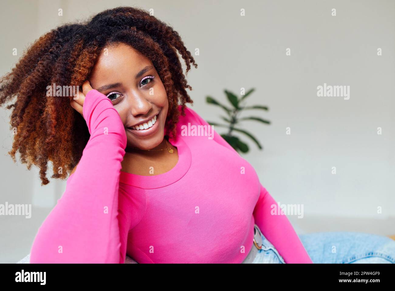 Die afroamerikanische Millennials trägt rosa, zufrieden und ruhig, sitzt auf dem bequemen Sofa im gemütlichen, modernen Wohnzimmer und hält die Hand im Gesicht. Stockfoto