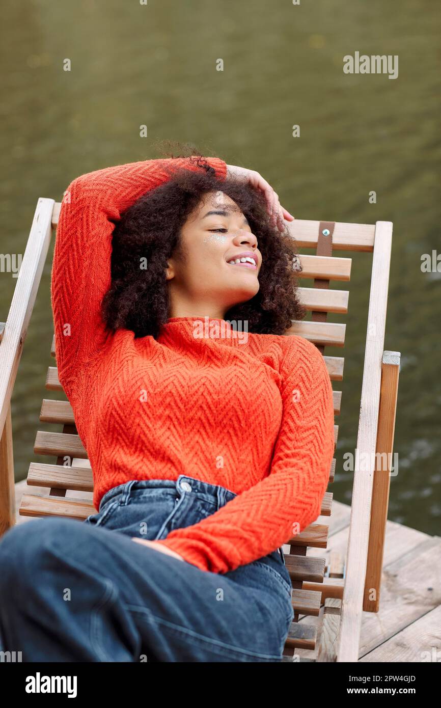 Junge, fröhliche, entspannte afroamerikanische Frau mit lockigem Haar in orangefarbenem, warmem Pullover, die sich im Herbst auf einem Liegestuhl in der Nähe eines Teichs oder Flusses ausruht Stockfoto