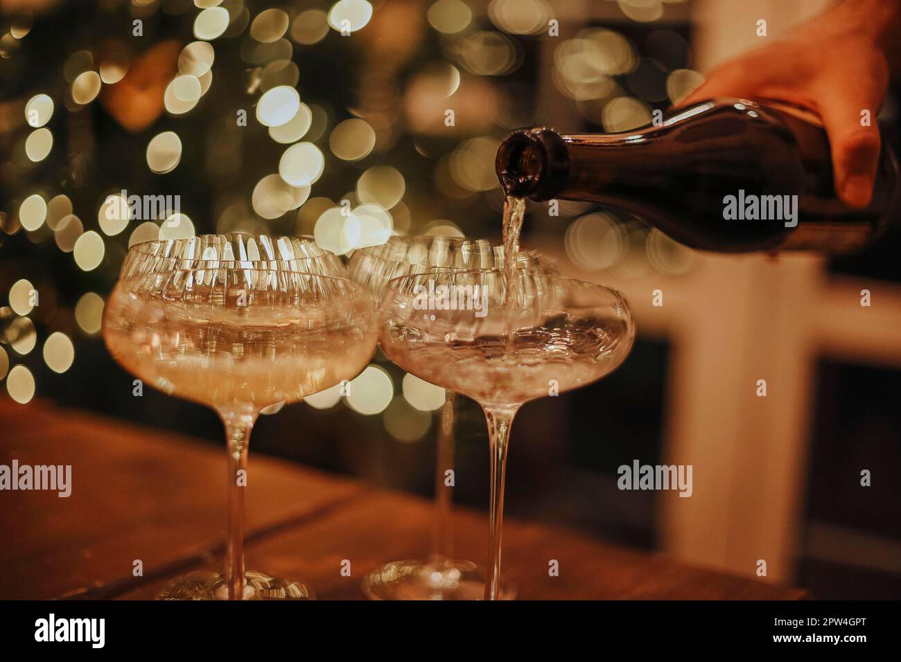 Silvesterfeier. Mann, der Champagner in Gläser gießt, steht auf dem Tisch mit festlichem Weihnachtsessen, Kerzen und Geschenkverpackungen gegen verschwommenes Bild Stockfoto