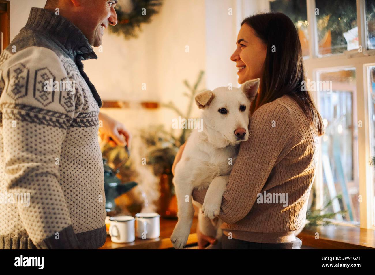 Ein junges glückliches Paar, Mann und Frau, halten einen süßen weißen Hund in der Hand und umarmen sich, während sie in der Küche stehen, dekoriert für Weihnachten, nette Familie mit Hündchen Stockfoto
