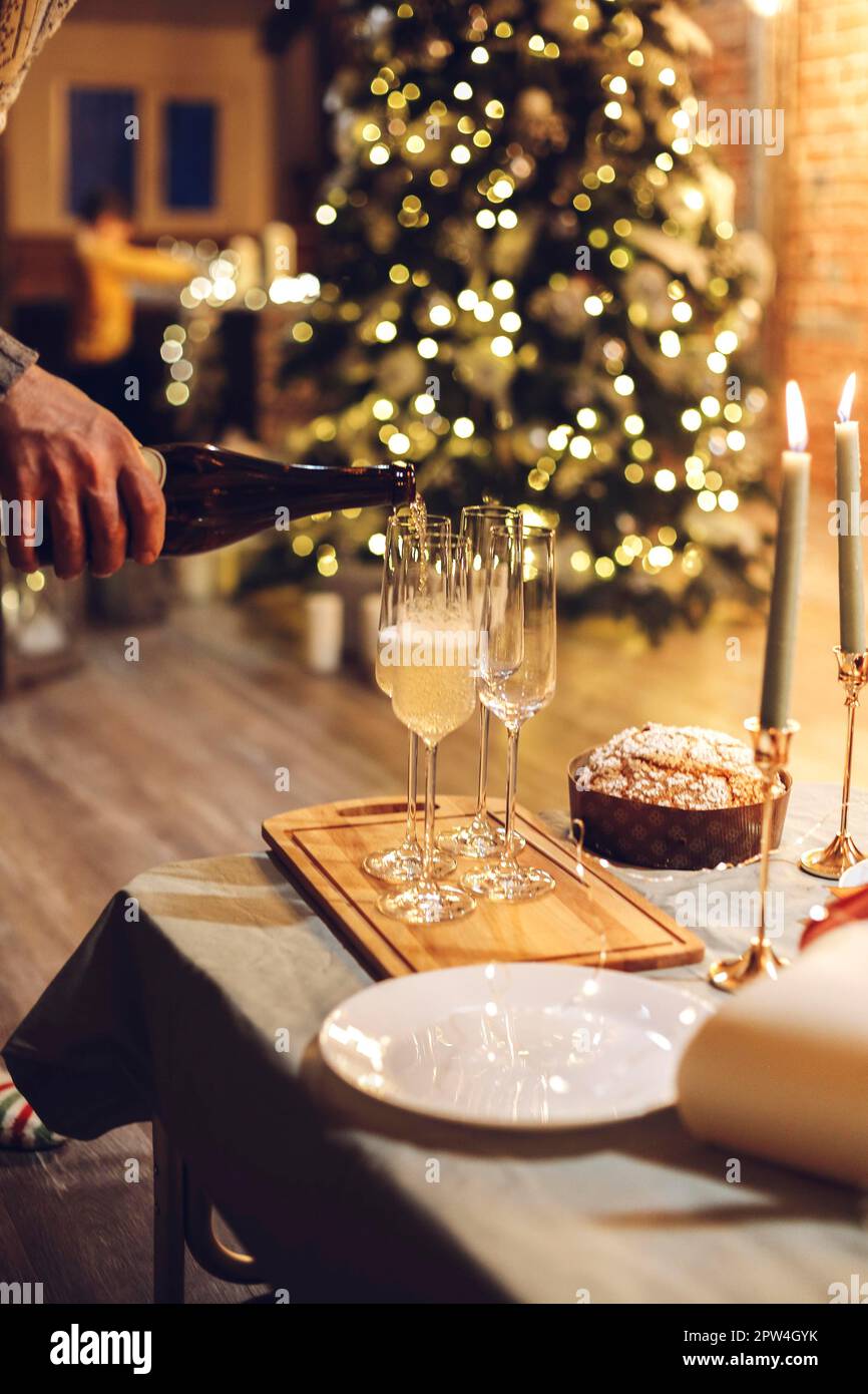 Silvesterfeier. Mann, der Champagner in Gläser gießt, steht auf dem Tisch mit festlichem Weihnachtsessen, Kerzen und Geschenkverpackungen gegen verschwommenes Bild Stockfoto