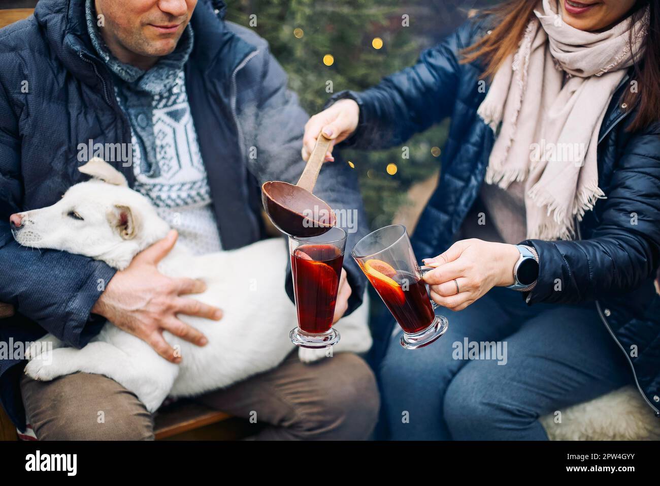 Nettes Paar mit niedlichem Hund, das draußen im Garten sitzt, umgeben von jungen Nadelbäumen, lächelt süß, während man miteinander kommuniziert Stockfoto