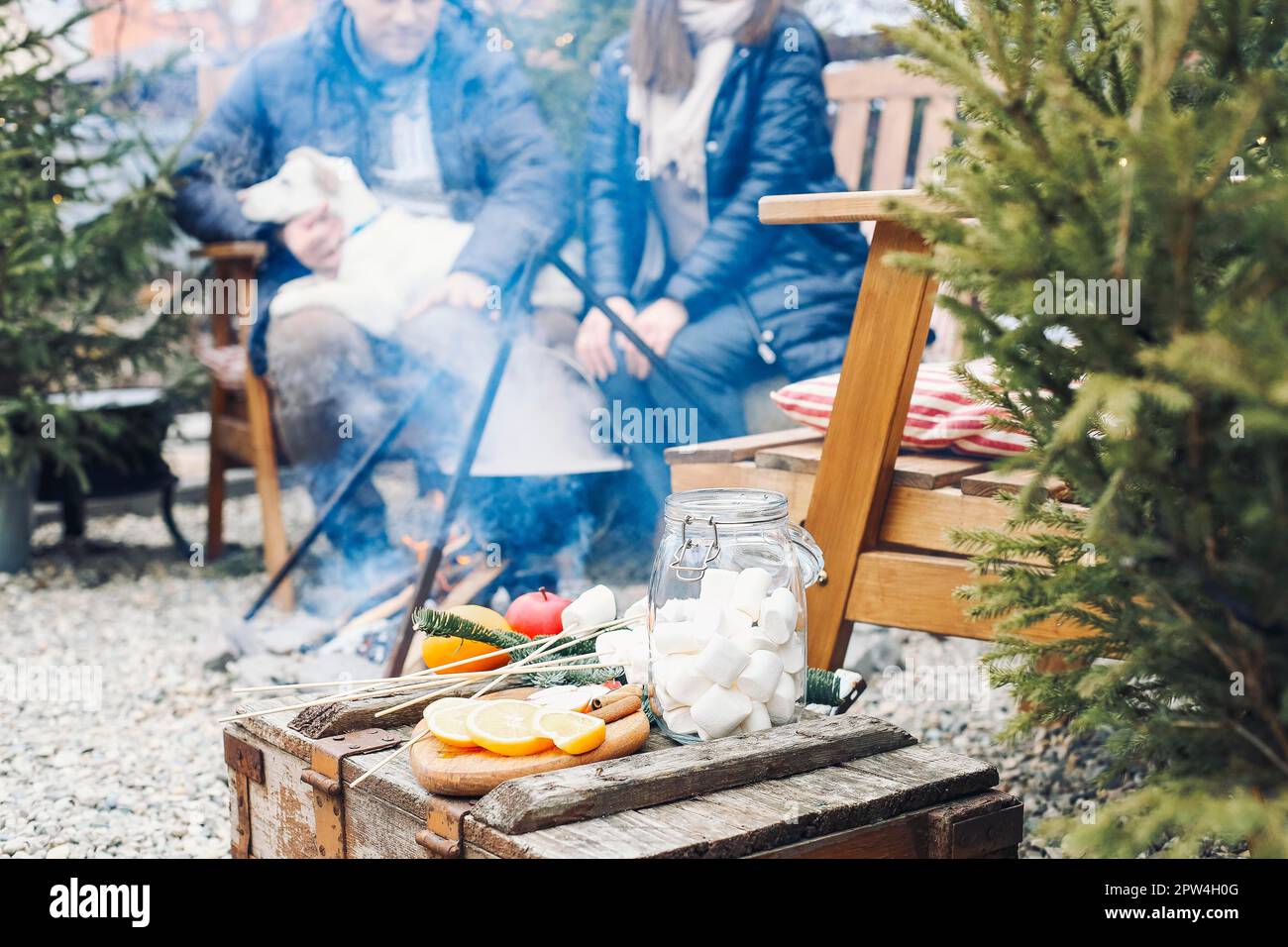 Nettes Paar mit niedlichem Hund, das draußen im Garten sitzt, umgeben von jungen Nadelbäumen, lächelt süß, während man miteinander kommuniziert Stockfoto