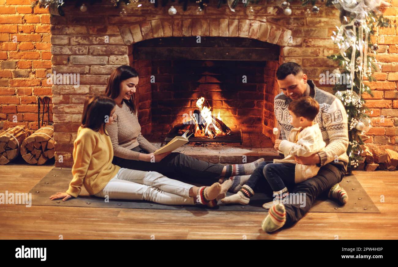 Glückliche europäische Familie mit Kindern, die am Kamin auf dem Boden sitzen, lesen und über dem Feuer marshallow braten, junge liebevolle Eltern mit zwei kleinen Kindern Stockfoto