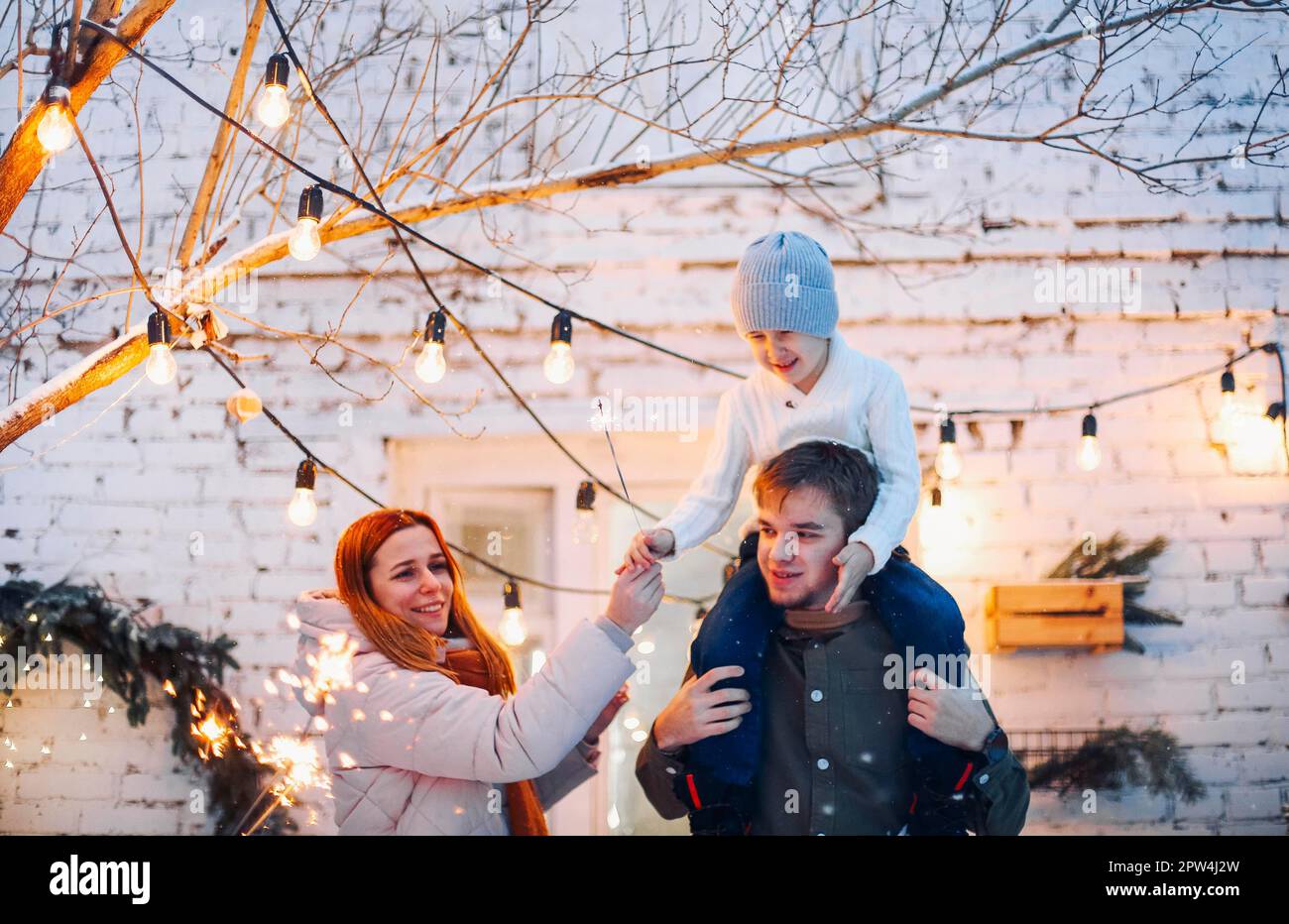 Eine glückliche Familie mit kindlichem Gefühl, während sie vor einem Ziegelhaus steht, das mit Tannenbäumen und wunderschönen Weihnachtslichtern dekoriert ist, Eltern Stockfoto