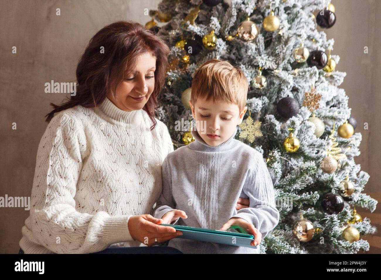 Großmutter und kleiner Enkel mit digitalem Tablet, die zu Hause neben dem wunderschön dekorierten Weihnachtsbaum sitzen und das Kind Zeit mit Oma verbringt Stockfoto