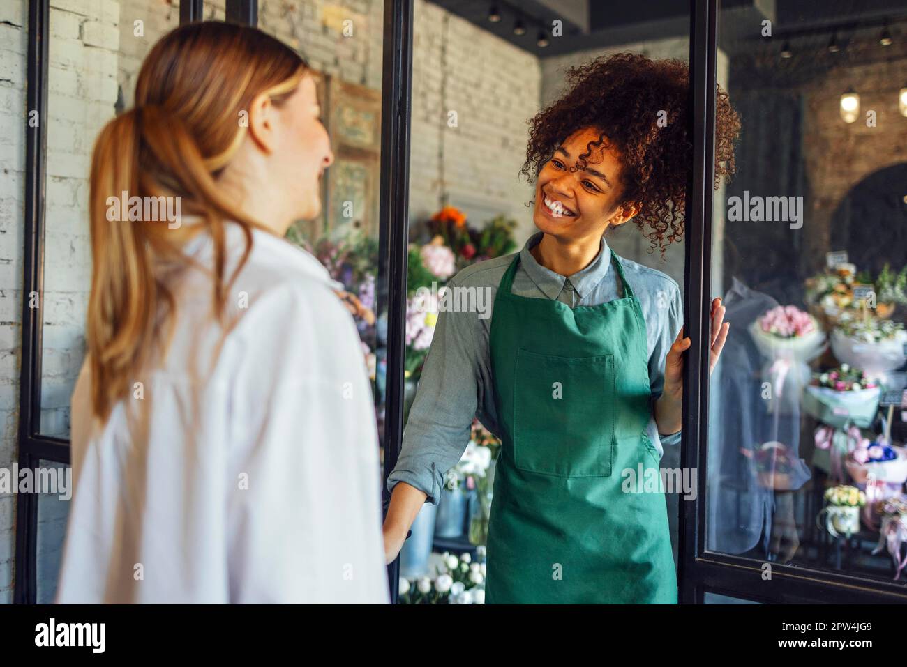 Eine glückliche schwarze Unternehmerin, die im Fabrikladen frische Blumen an Kunden verkauft. Junge Blonde kauft einen frischen Blumenstrauß vom Blumenhändler. Lächelnd Stockfoto