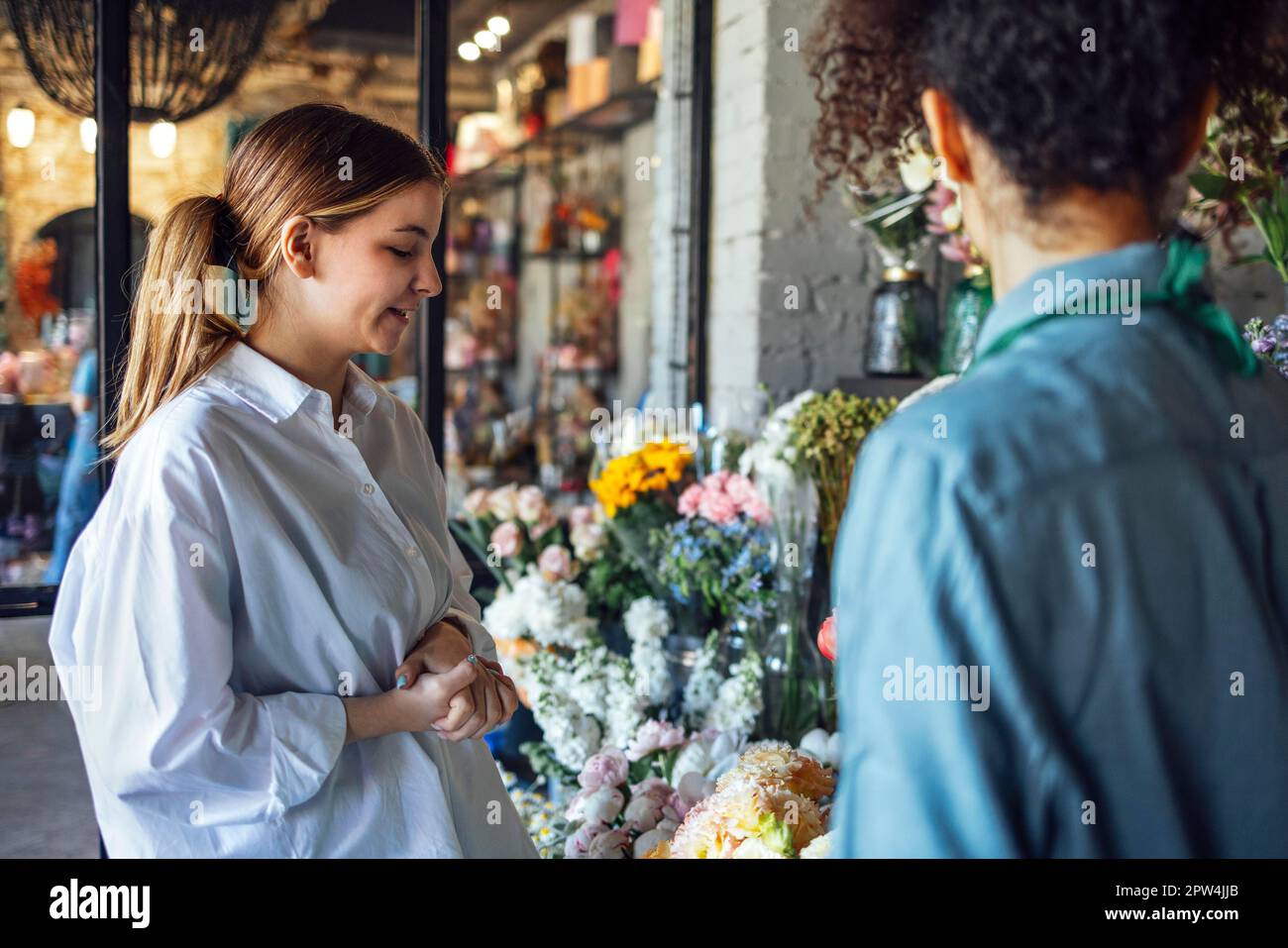 Eine glückliche schwarze Unternehmerin, die im Fabrikladen frische Blumen an Kunden verkauft. Junge Blonde kauft einen frischen Blumenstrauß vom Blumenhändler. Lächelnd Stockfoto
