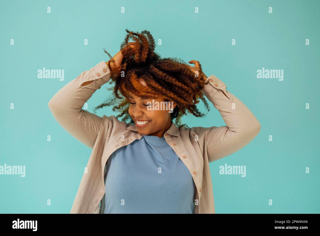 Positive afroamerikanische weibliche Model lächelt an der Kamera und berührt sauberes lockiges Haar nach der Hygienebehandlung auf blauem Hintergrund Stockfoto